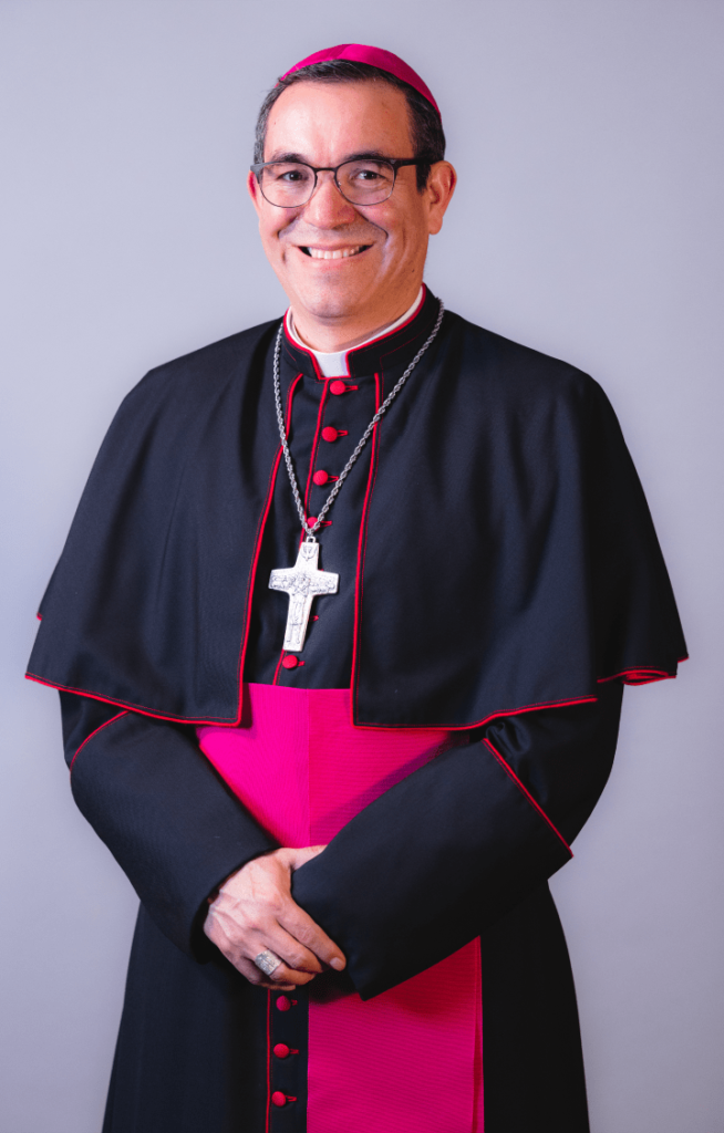 A clergymember wearing traditional black and red vestments, including a cross necklace and a biretta, stands smiling with hands clasped.