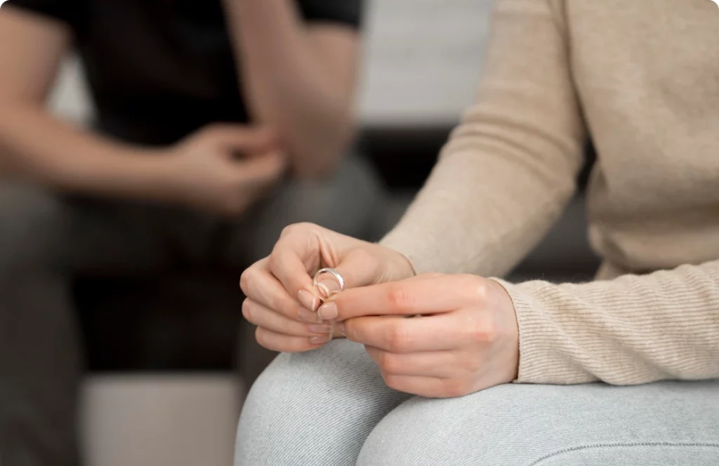 A person sitting holds a ring in their hands, with another person in the background sitting with arms crossed. Both individuals are dressed in casual attire, with a focus on the hands and ring.