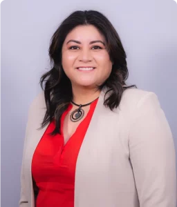 A person with dark hair wearing a beige blazer and a red top is smiling against a light gray background.