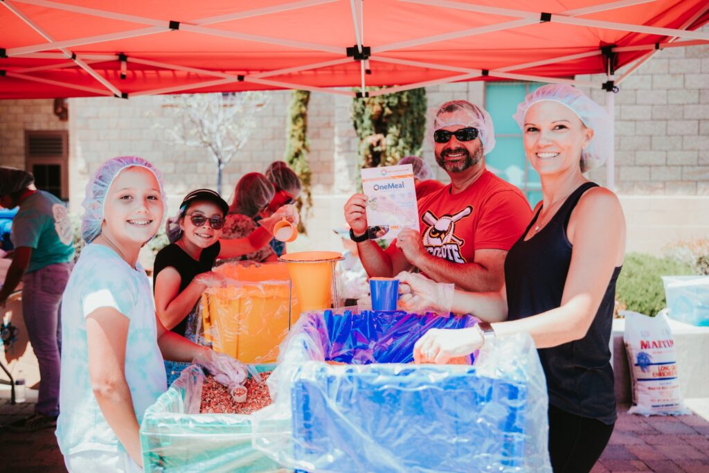 Las personas bajo un toldo rojo llenan bolsas con alimentos procedentes de grandes contenedores. Llevan redecillas para el pelo y guantes, y sonríen a la cámara. Una persona sostiene un paquete de comida con la etiqueta «OneMeal».