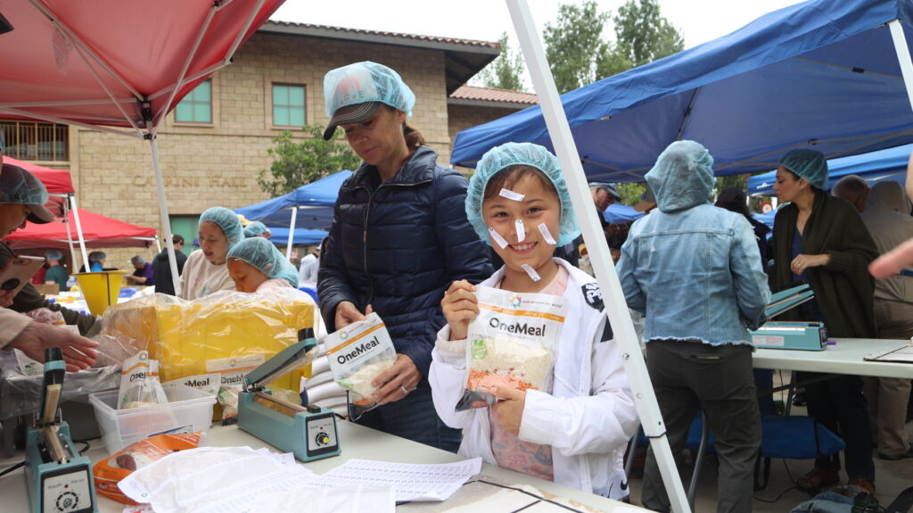 Un grupo de personas con gorros y chaquetas están empaquetando comida en un evento al aire libre. Una persona en primer plano sonríe y sostiene una bolsa con la etiqueta «OneMeal» bajo una carpa roja.
