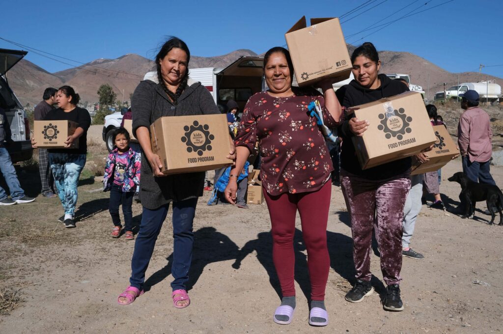 Personas transportando cajas de alimentos con la etiqueta «Kids Around the World» (Niños de todo el mundo) en una zona rural con montañas al fondo. También hay un niño y un perro.