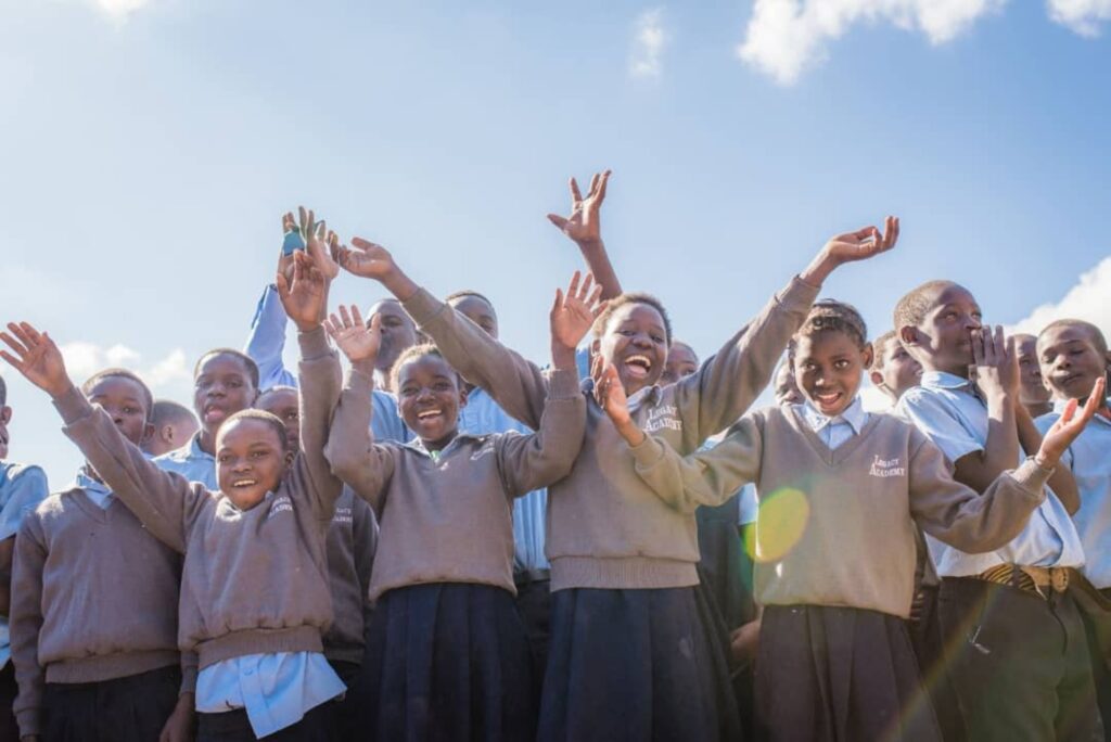 Un grupo de escolares con uniforme están de pie al aire libre, sonriendo y levantando los brazos contra un cielo azul claro.