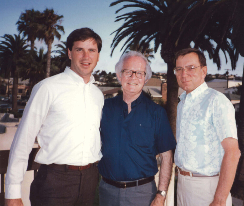 Three people stand together outdoors, smiling. The person on the left wears a white shirt, the center person wears a dark shirt with glasses, and the right person wears a patterned shirt. They are surrounded by palm trees and a clear sky.